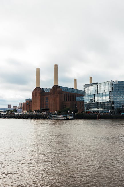 A view across a river shows a large, historic brick building with four tall, cylindrical chimneys extending upward, situated on the riverbank. Adjacent to the brick structure is a modern glass-fronted office building with reflective windows, creating a contrast between old and new architecture. The river in the foreground has a slightly rippled surface, with several boats docked along the shoreline. The sky above is overcast with thick, gray clouds, casting diffuse light over the scene. The building complex, including the chimney-stack factory and the contemporary glass office, appears as part of an urban riverside environment, potentially involved in industrial or commercial activities, which may relate to the broader context of alternative waste handling or localised rubbish collection, as offered by private waste management services like Rubbish Collection Battersea.