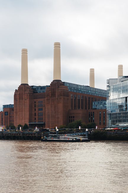 A large, historic power station building located along the riverbank, constructed from red-brick material with a textured surface, featuring tall, narrow vertical windows and four prominent, cream-colored chimney stacks extending upward from the structure. Adjacent to the power station are modern glass-fronted office buildings with reflective surfaces that mirror the overcast sky. In the foreground, a section of the river with brownish water is visible, along with a small boat moored near the water's edge. The overall scene suggests an urban riverside environment, with the power station and nearby buildings situated on a paved area or dock. The lighting is diffuse, typical of cloudy weather, providing soft illumination across the scene. This setting aligns with the context of waste management or rubbish removal services operating in historic or industrial urban areas, emphasizing the importance of alternative disposal methods like private rubbish collection for such sites. Rubbish Collection Battersea might facilitate cleanup or waste removal in these environments, supporting efficient rubbish handling outside of traditional municipal services.