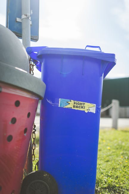A tall, upright outdoor blue rubbish bin with a matching lid sits on a paved area beside a grassy verge, prominently displaying a yellow and blue sticker that reads 'FIGHT BACK!'. To the left, part of a red wheelie bin with black circular ventilation holes and a silver lid is visible, with a chain attached to the bin's side. In the background, a clear sky with some light cloud cover and a portion of a vehicle or sign structure can be seen, indicating a typical waste collection setting. The scene is well-lit with natural daylight, suggesting a bright, possibly sunny day. The overall environment suggests an outdoor urban or suburban location where private rubbish collection services, such as those provided by Rubbish Collection Battersea, are used for waste management and collection alternatives outside of local authority provisions. The arrangement and condition of the bins imply that waste is prepared for disposal or collection, supporting the theme of independent rubbish removal.
